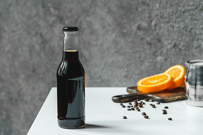 selective focus of cold brew coffee with ice in bottle near orange slices on chopping board and coffee beans on white table