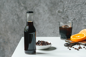 selective focus of cold brew coffee with ice in bottle and glass near orange slices on chopping board and coffee beans on white table