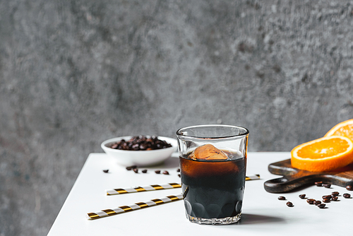 selective focus of cold brew coffee with ice in glass near orange slices on chopping board,  straws, coffee beans on white table