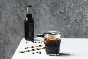 selective focus of cold brew coffee with ice in glass and bottle near  straws and coffee beans on white table