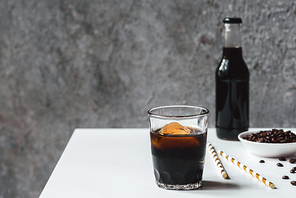 selective focus of cold brew coffee with ice in glass and bottle near  straws and coffee beans on white table