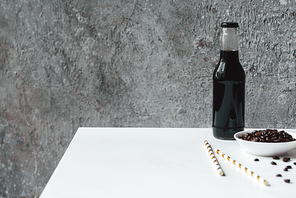 selective focus of cold brew coffee with ice in bottle near  straws and coffee beans on white table