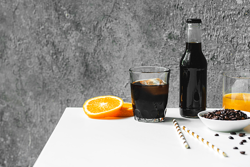 selective focus of cold brew coffee with ice in glass and bottle near orange juice,  straws and coffee beans on white table
