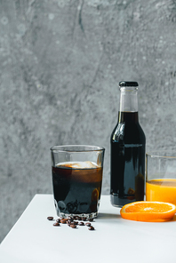 cold brew coffee with ice in glass and bottle near orange juice and coffee beans on white table