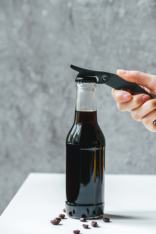 cropped view of woman opening cold brew coffee in bottle with opener on white table with coffee beans