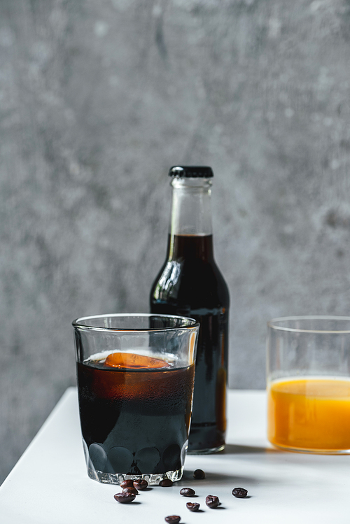 selective focus of cold brew coffee with ice in glass and bottle near orange juice and coffee beans on white table