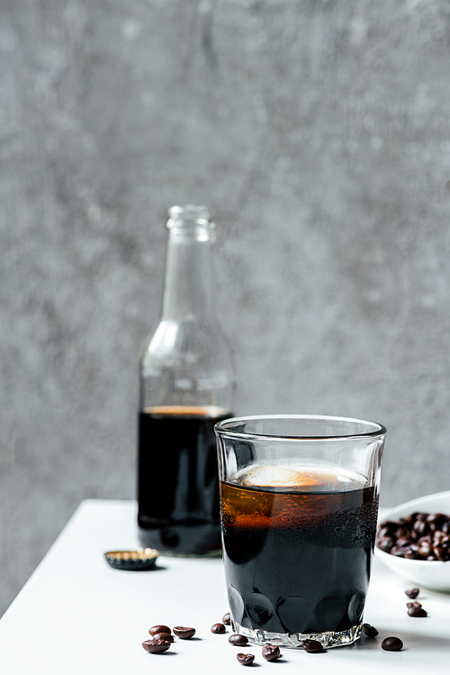selective focus of cold brew coffee with ice in glass near bottle and coffee beans on white table