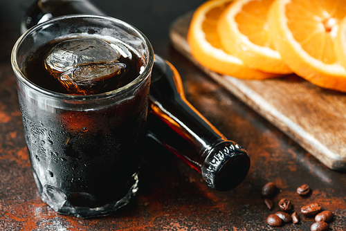 selective focus of cold brew coffee with ice in glass and bottle near orange slices and coffee beans on rusty surface