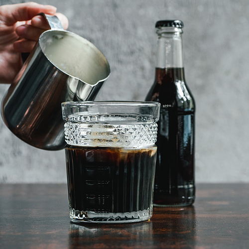 cropped view of woman pouring milk in cold brew coffee in glass near bottle on wooden table
