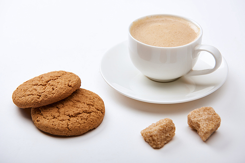 tasty coffee with foam in cup on saucer near brown sugar and cookies on white background