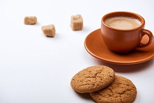 delicious coffee with foam in brown cup on saucer with brown sugar and cookies on white background