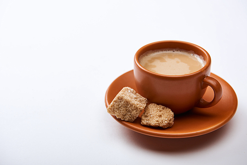 tasty coffee with foam in cup on saucer near brown sugar on white background with copy space