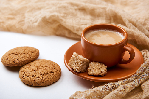 delicious coffee with foam in cup on saucer with brown sugar near cloth and cookies on white background