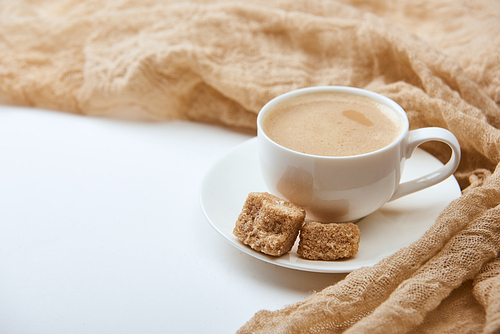 delicious coffee with foam in white cup on saucer with brown sugar near cloth on white background