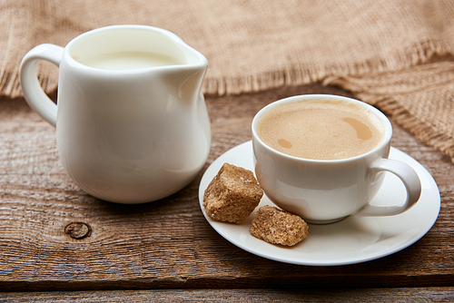 delicious coffee with foam in cup on saucer with brown sugar near sackcloth and jug milk on wooden background