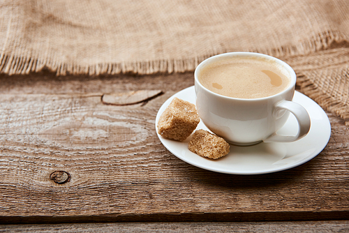 delicious coffee with foam in cup on saucer with brown sugar near sackcloth on wooden background