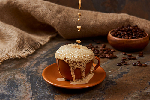 pouring hot coffee into cup with foam on marble surface near sackcloth and beans