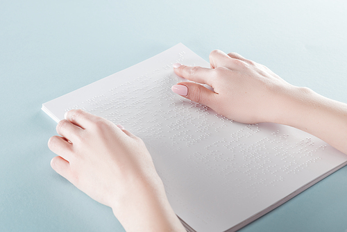 cropped view of young woman reading braille text on paper isolated on blue