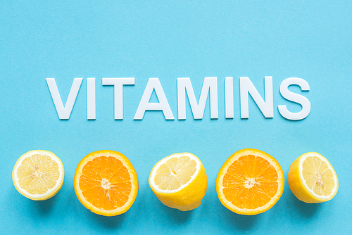 top view of ripe orange, lemon halves and word vitamins on blue background