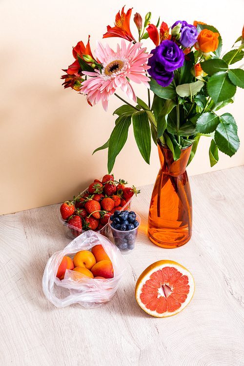 floral and fruit composition with bouquet in vase, berries, grapefruit and apricots on wooden surface on beige background