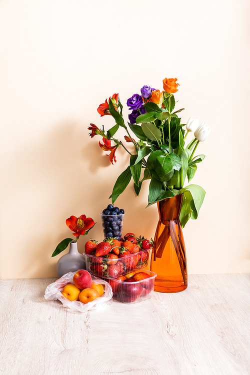 floral and fruit composition with bouquet in vase and summer fruits on wooden surface on beige background