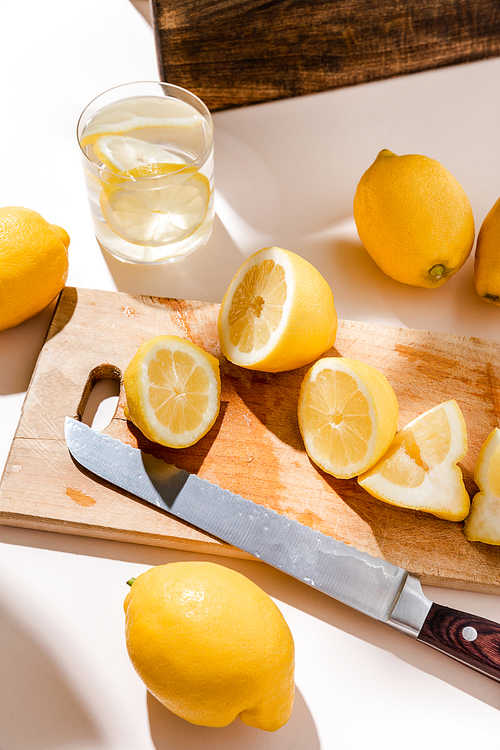 whole and cutted lemons on wooden board with knife and glass of water on grey table