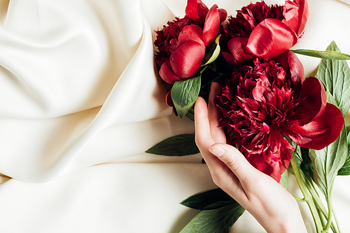 top view of female hand and bouquet of red peonies on white cloth