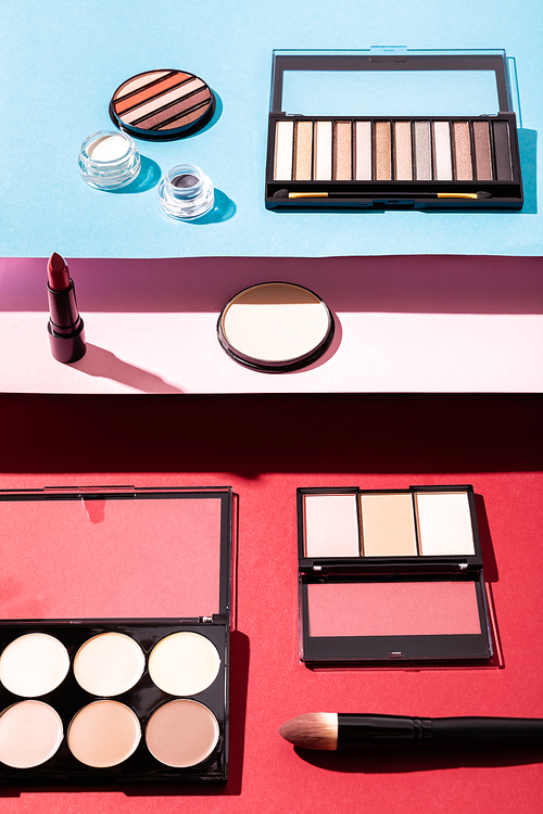 blush and eye shadow palettes near cosmetic brushes, lipstick and face powder on crimson, pink and blue