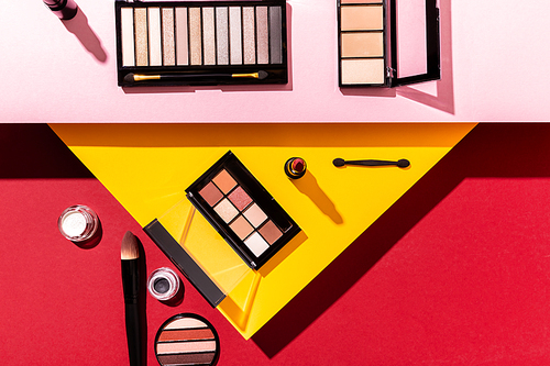 top view of eye shadow palettes and cosmetic brushes near lipstick on crimson, pink and yellow