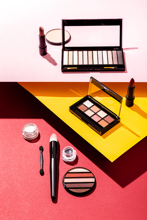 eye shadow palettes and cosmetic brushes near lipsticks and face powder on crimson, pink and yellow