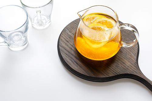 hot tea in teapot with lemon and ginger on wooden board near empty cups on white background