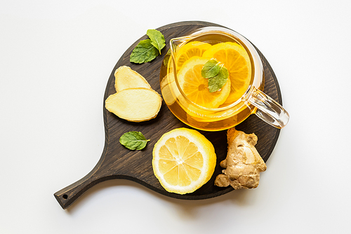top view of hot tea in teapot served on wooden board with ginger root, lemon and mint on white background