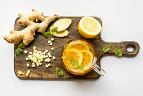 top view of hot tea near ginger root, lemon and mint on wooden cutting board on white background