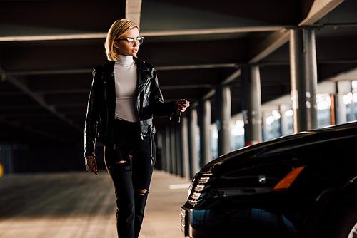 pretty young blonde woman in glasses walking in parking with key near car
