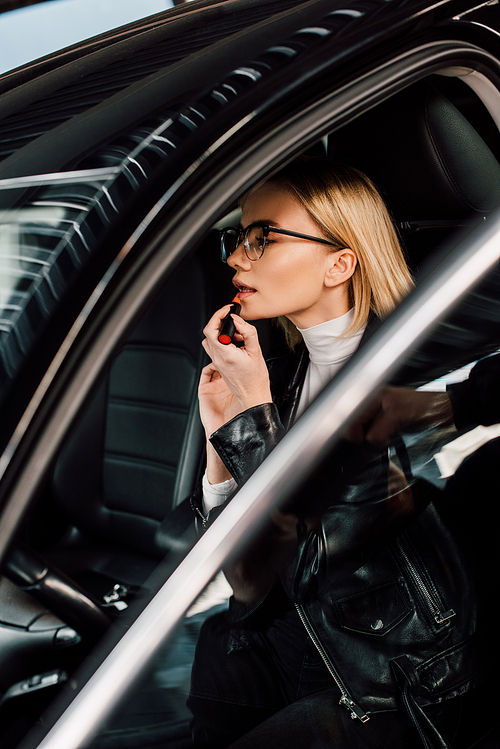 attractive blonde girl in glasses applying lipstick in car