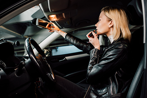 low angle view of attractive blonde girl applying lipstick and touching mirror in car