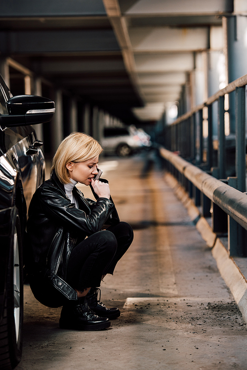 worried blonde girl talking on smartphone while sitting near car