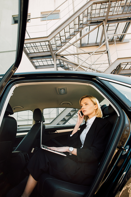 low angle view of worried blonde woman talking on smartphone while sitting with laptop in car