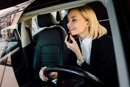 cheerful blonde young woman applying lipstick in car