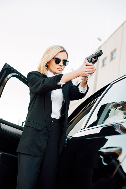 dangerous blonde young woman in sunglasses holding gun near car
