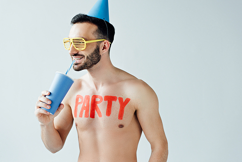 shirtless man in party hat with inscription on body drinking beverage isolated on grey