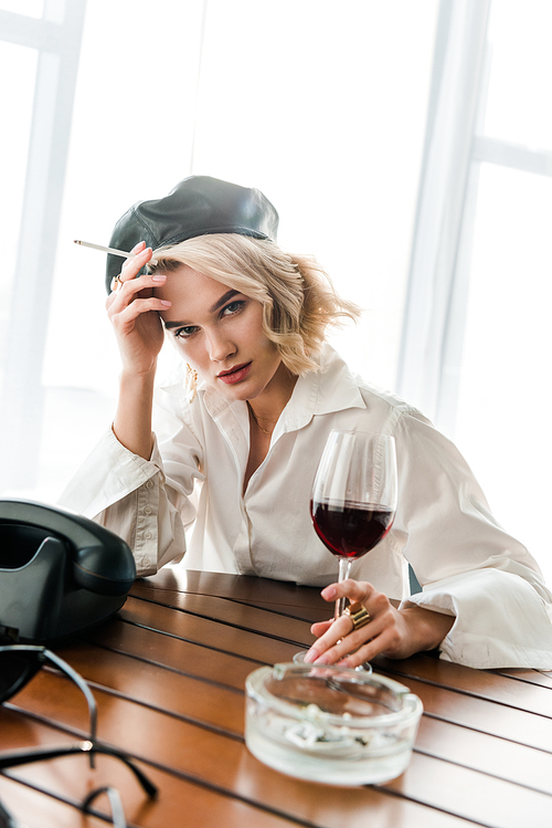 elegant blonde woman in black beret and white shirt  holding cigarette and glass with red wine