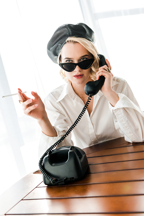 elegant blonde woman in black beret and sunglasses smoking cigarette while talking on retro phone