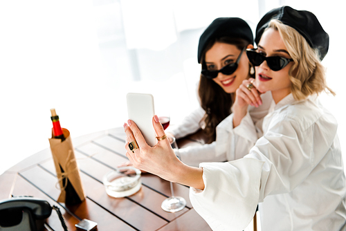 selective focus of elegant brunette and blonde women in black berets and sunglasses drinking red wine and taking selfie