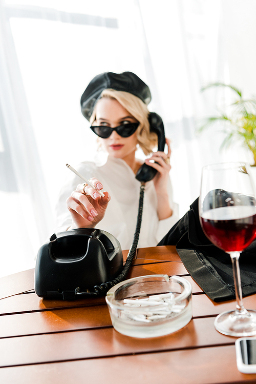 selective focus of elegant blonde woman in black beret and sunglasses smoking and talking retro phone near glass with red wine