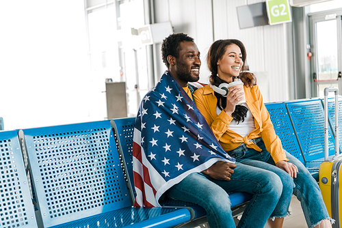 happy african american couple sitting with american flag and coffee to go in departure lounge in airport