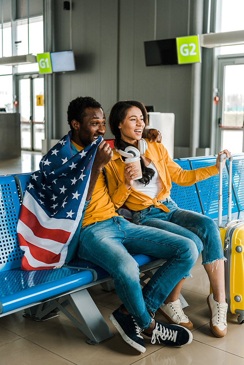 happy african american couple sitting with american flag and luggage in departure lounge in airport