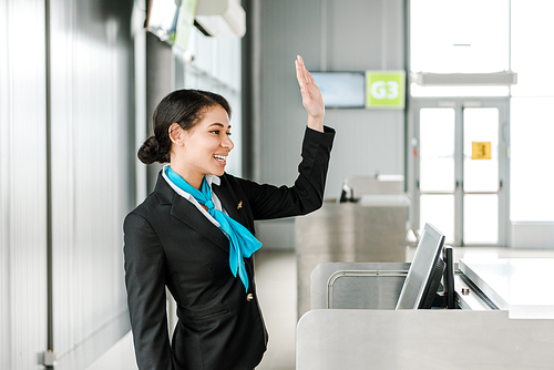 smiling african american airport staff in uniform standing at check in desk and waving hand
