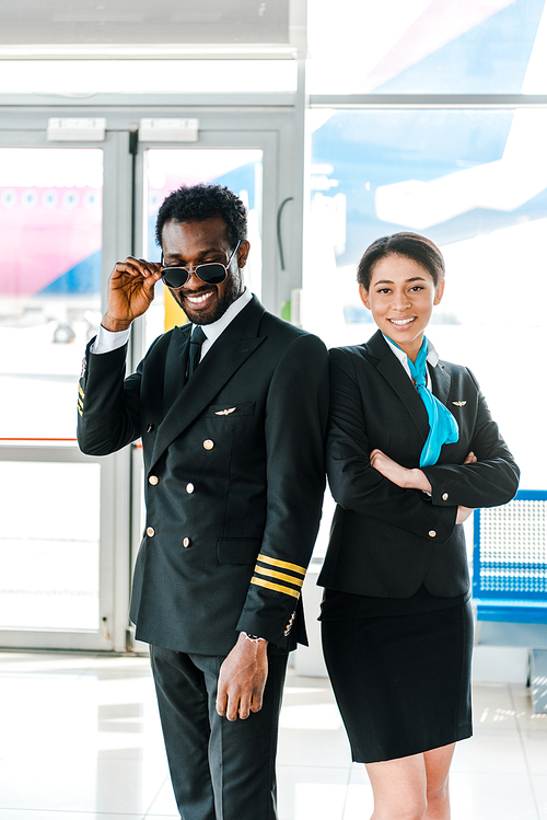 african american pilot in sunglasses and stewardess with crossed arms posing together in airport