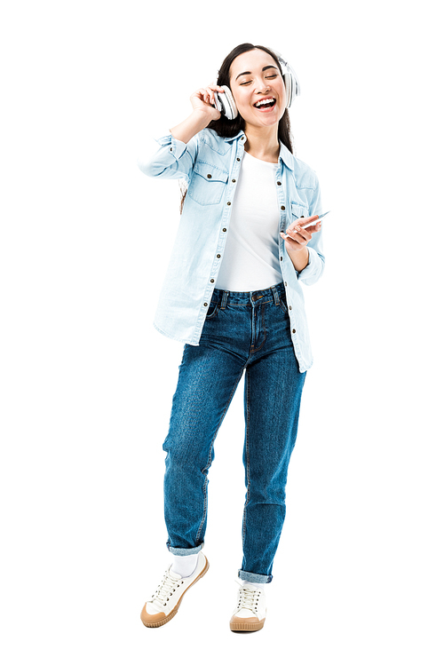 attractive and smiling asian woman in denim shirt listening music and holding smartphone isolated on white